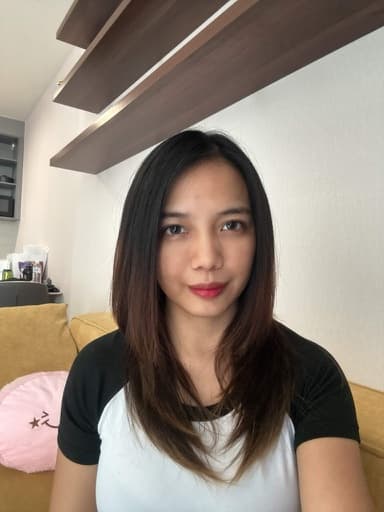 Anne sitting on a yellow couch in front of a beige wall with floating wooden shelves above. She has long dark hair and wears a white t-shirt with black raglan sleeves.