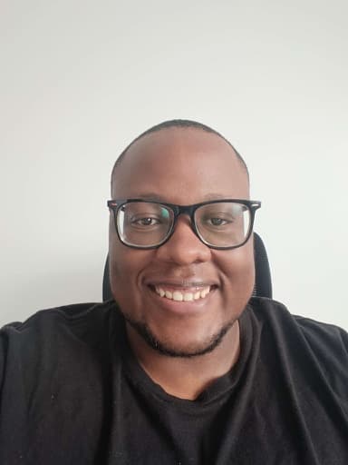 Hanningtone seated in an office chair against a plain white wall. He has a shaved head, dark-framed glasses, and a light beard, wearing a black t-shirt and smiling at the camera.