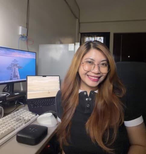 Mae at her desk with a monitor, laptop, and mechanical keyboard visible behind her. She wears glasses and a black collared top and is smiling.